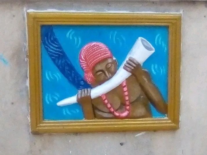 Framed, painted relief of a man in a red cap and coral beads blowing an ivory horn